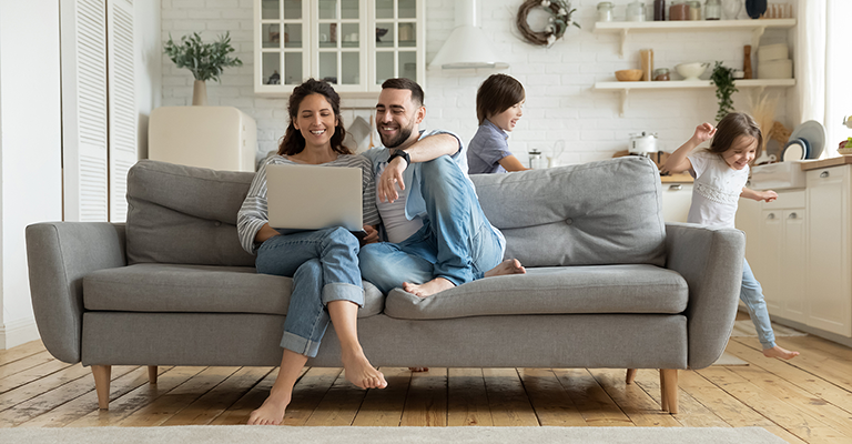 a family, with the parents looking at a laptop on the couch and kids playing